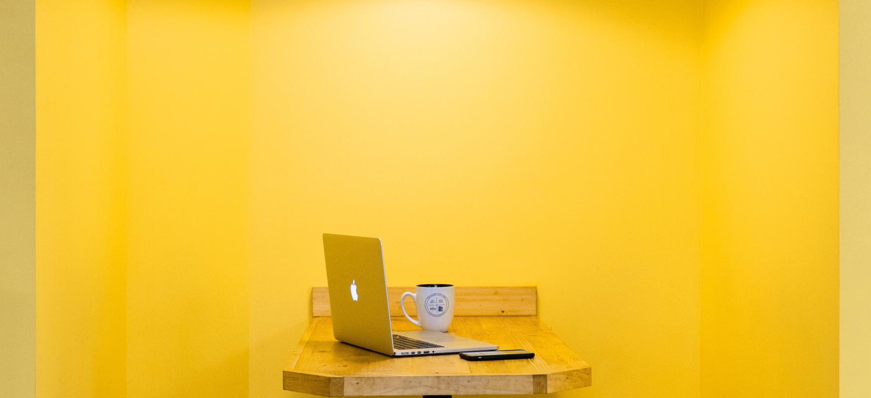 Booth with two benches and a table with a laptop on top. Yellow wall.