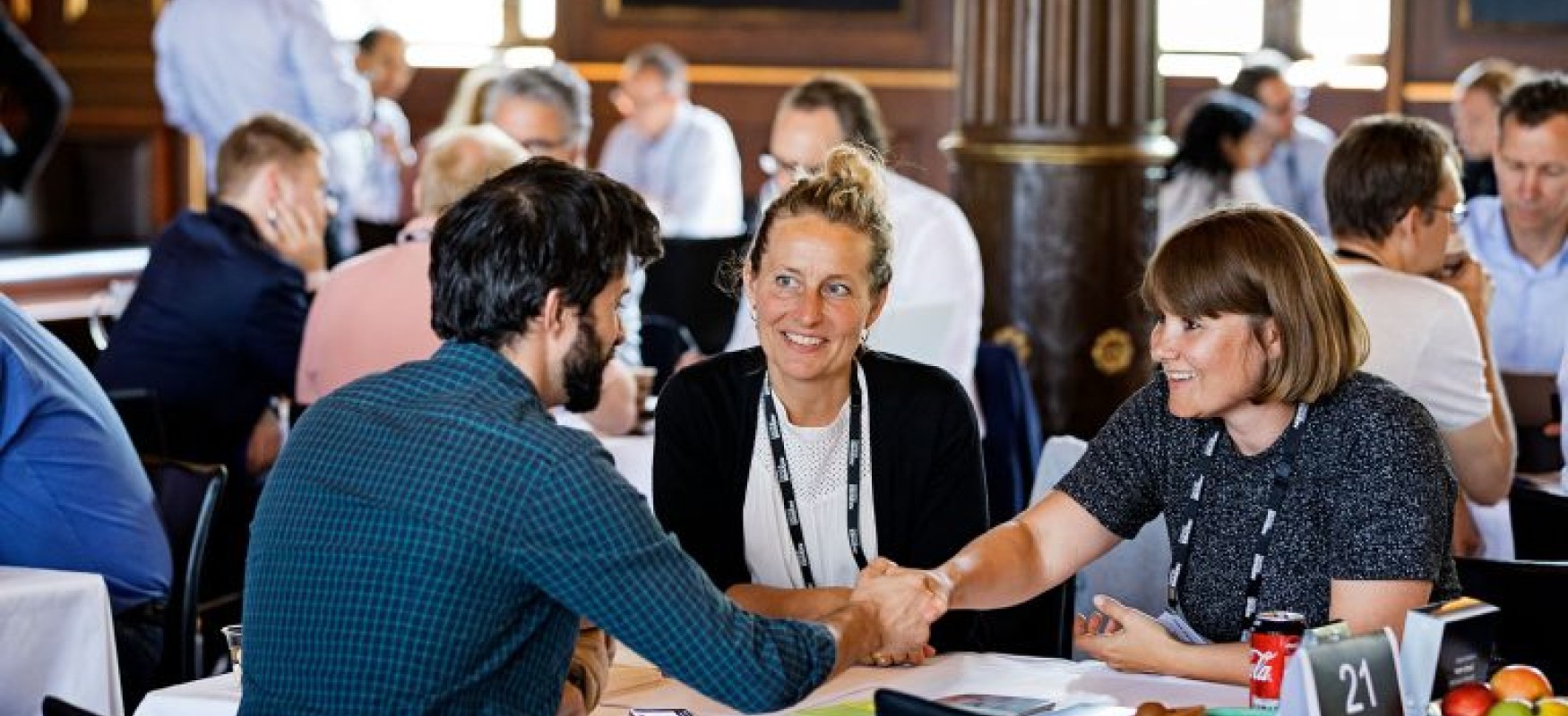 Two persons shaking hands with another person is watching while they all sit around a roundtable at the Nordic Impact Business Summit 2019.