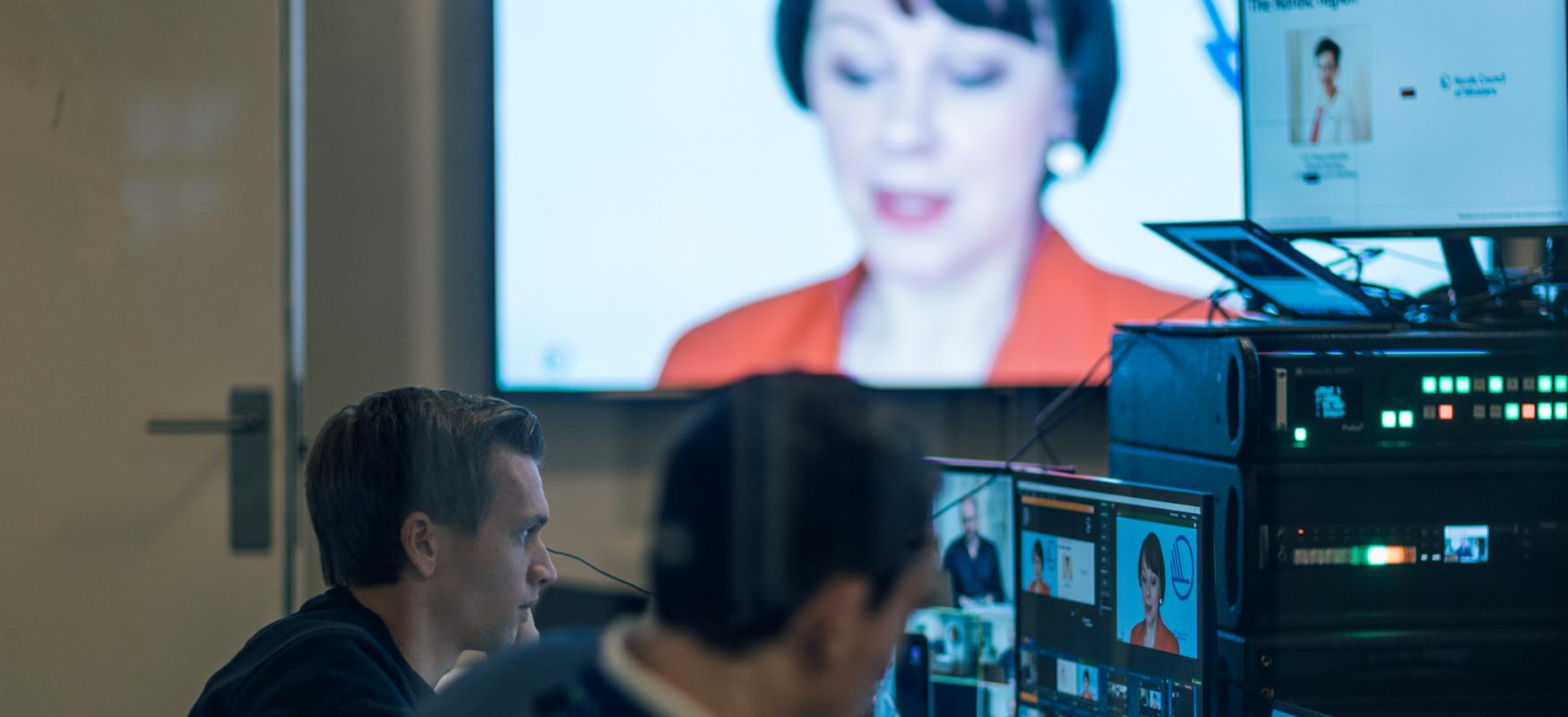 Two men sitting in front of computer screens overseeing a webinar. Paula Lehtomaki pictured on a big screen in the background.