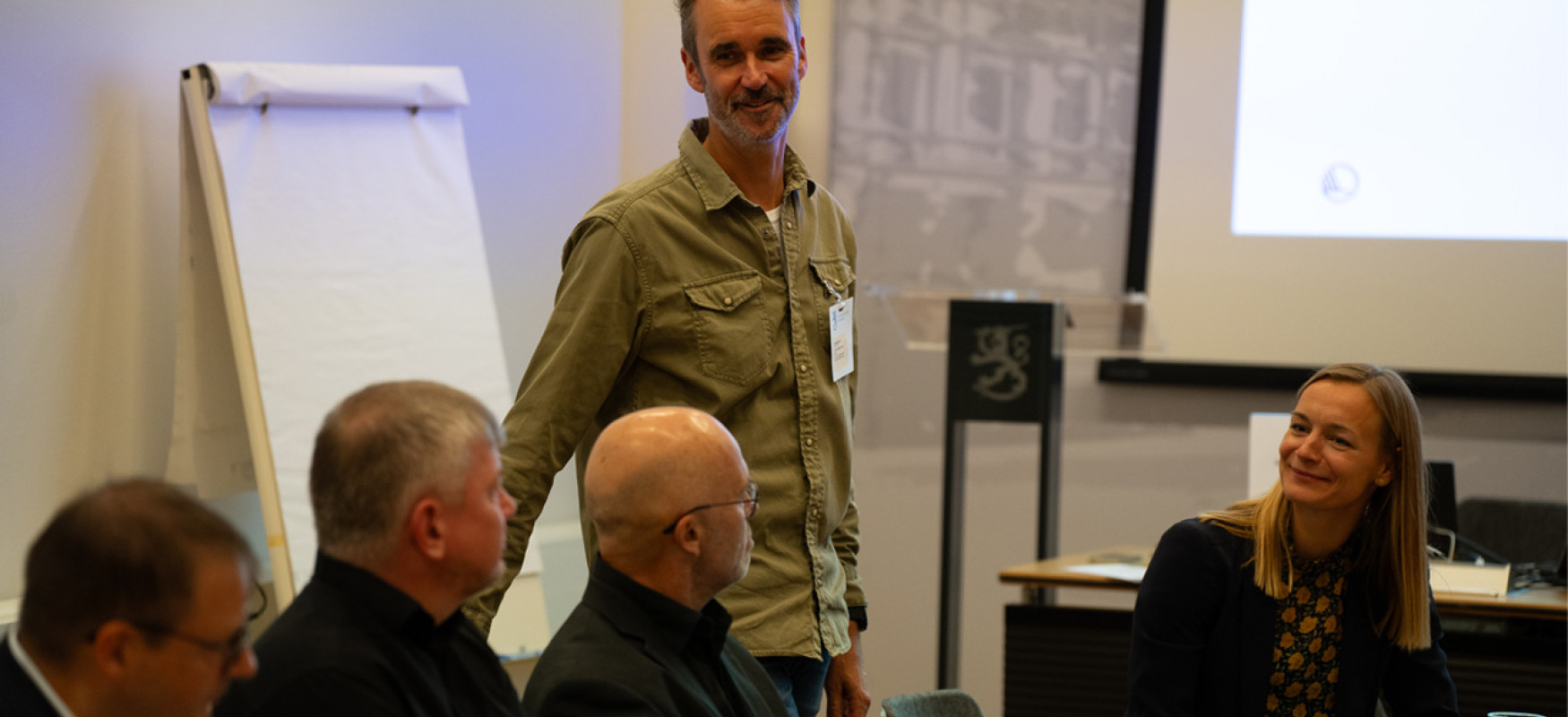 Man standing up and talking to an audience gathere round a table