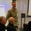 Man standing up and talking to an audience gathere round a table